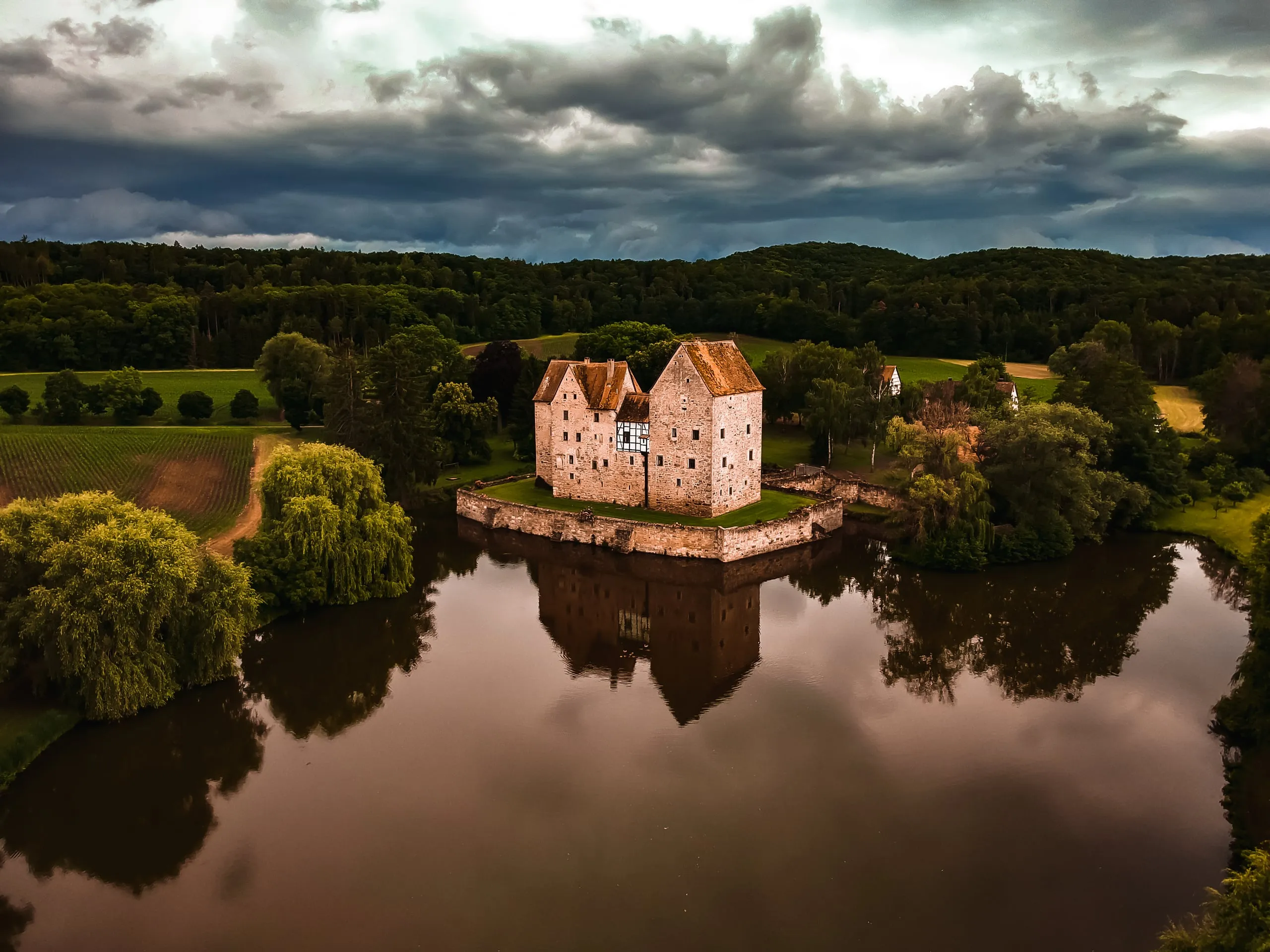 Vodní hrad Brennhausen na ostrůvku uprostřed rybníka, nad ním bouřková oblaka