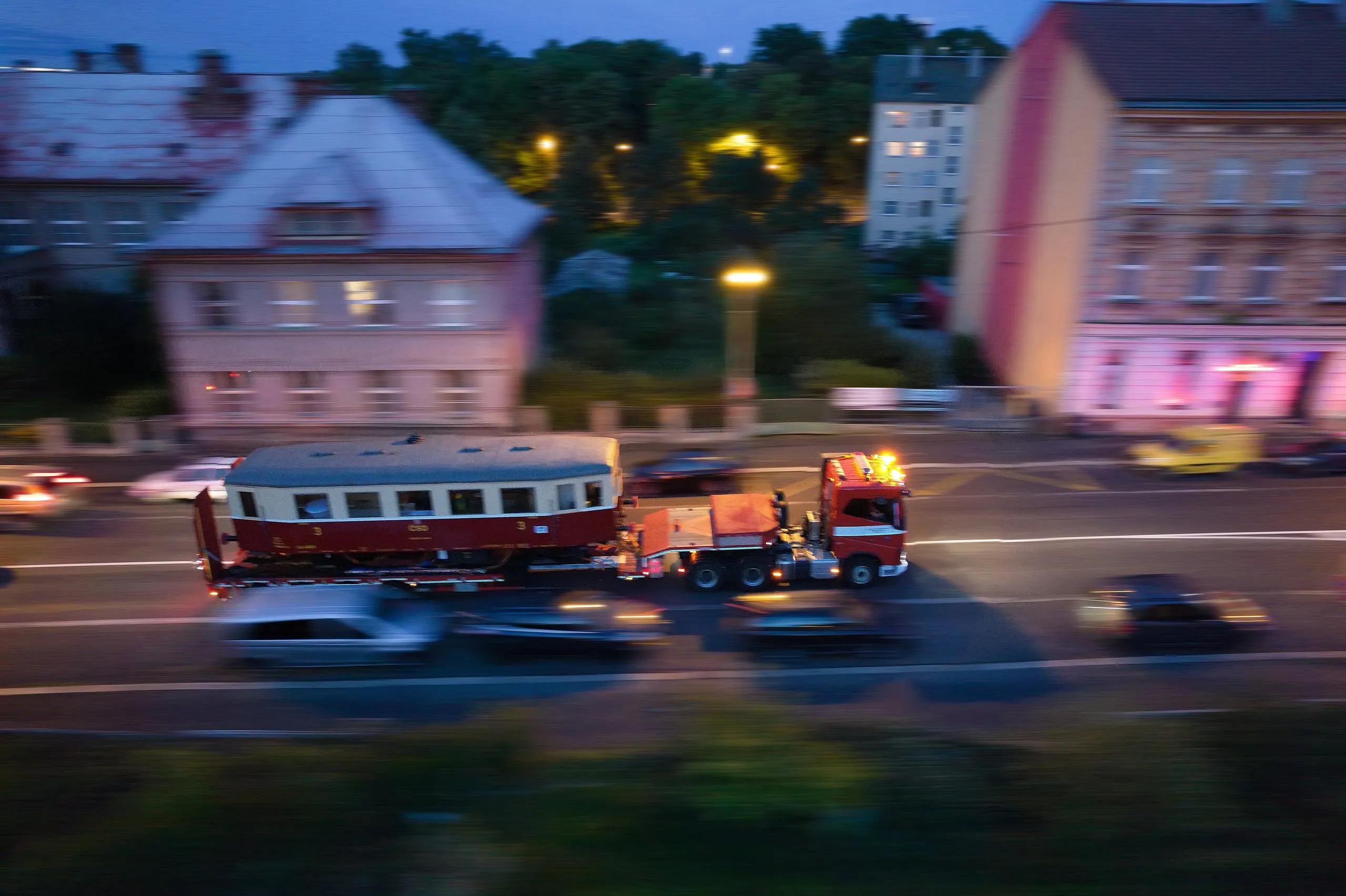 Drážní hasiči převážejí motorový vůz na podvalníku večerní ulicí
