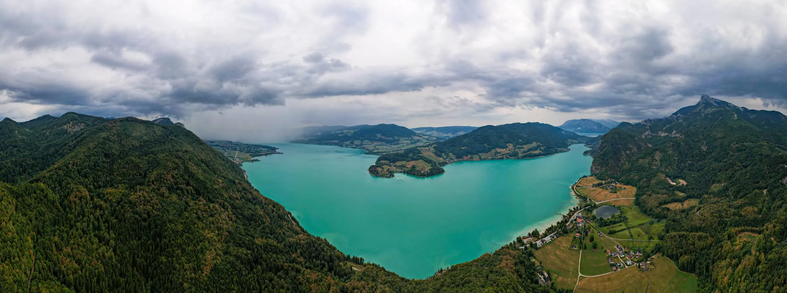 Panorama tyrkysového jezera Mondsee s poloostrovem pod zataženou oblohou