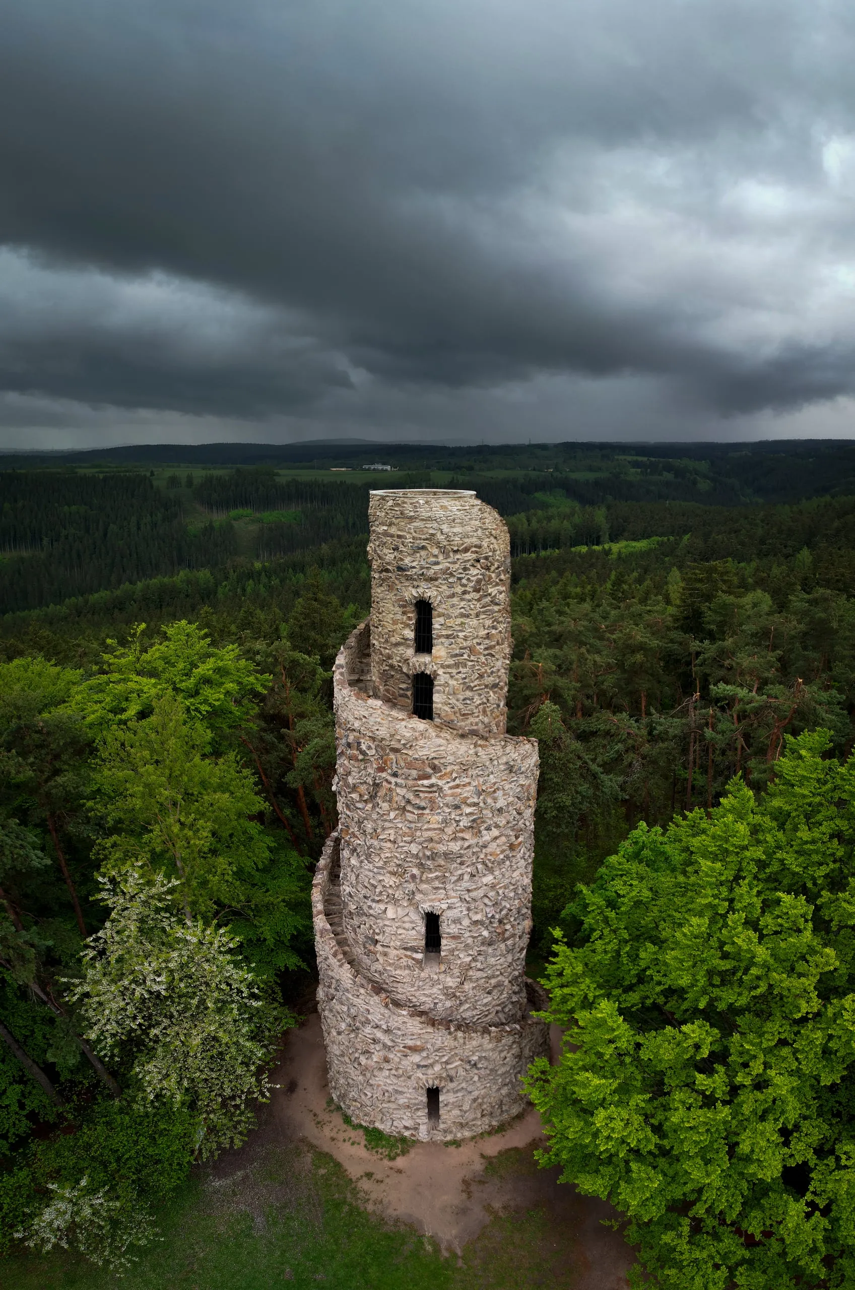 Kamenná rozhledna Krásno se spirálovým schodištěm nad lesy, nad ní bouřkové mraky